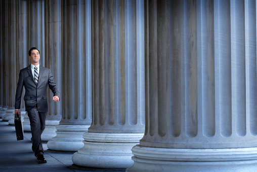 A businessman or a lawyer, carrying a briefcase, walking past a long row of columns.  The businessman is walking with purpose and confidence. He is placed on the far left side of the image leaving the right side open for copy.