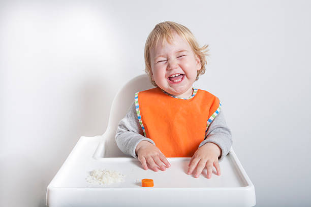 laughing in meal blonde caucasian baby seventeen month age orange bib grey sweater eating rice carrot on white high-chair laughing and smiling baby bib stock pictures, royalty-free photos & images