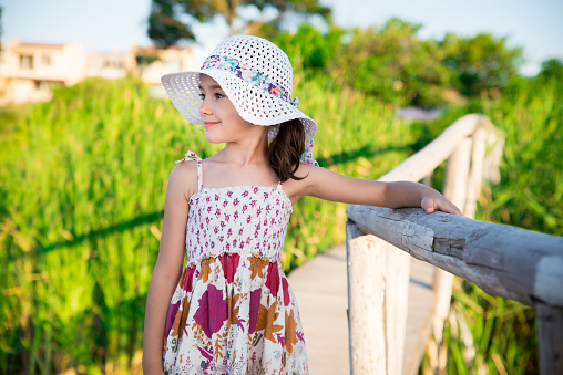 Candid lifestyle portrait in profile of beautiful smiled white Caucasian girl looking away from the camera, wearing white summer floppy hat and dress with fresh flower printed motives in orange and purple colours while holding to handrail of timber made wooden bridge in nature. Shot on Canon EOS, ISO 100.