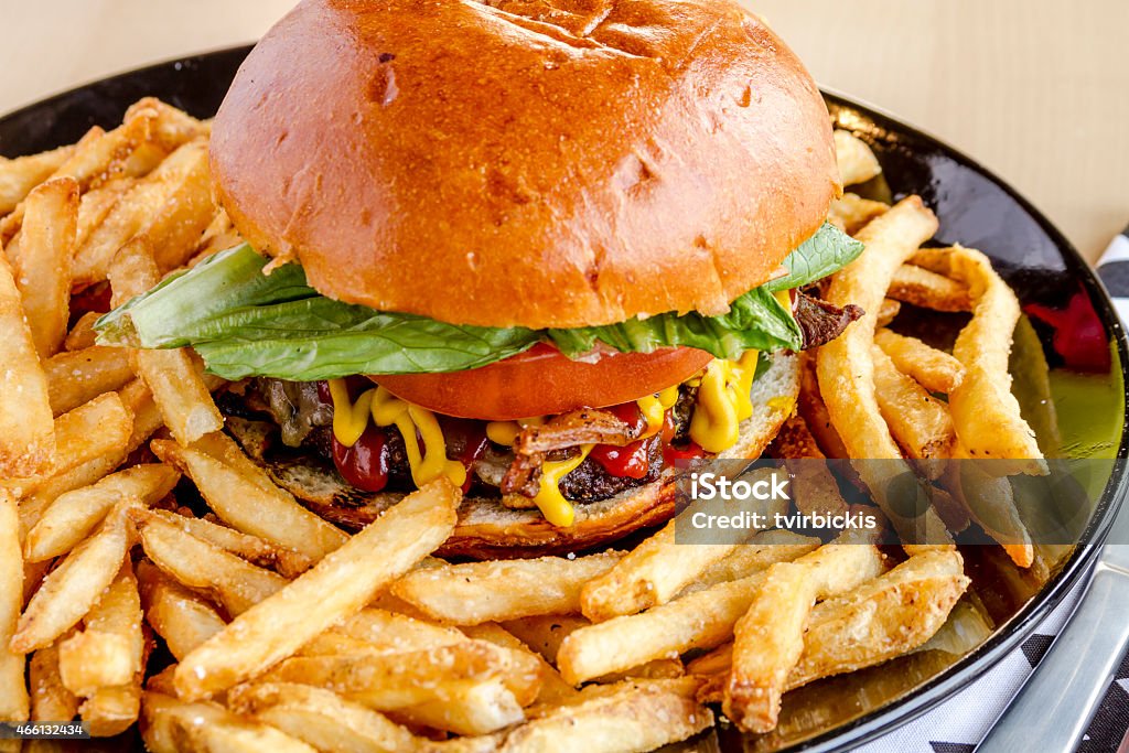 Gourmet Pub Hamburger and Fries Close up of gourmet pub hamburger with bacon on black plate with french fries sitting on wooden table 2015 Stock Photo