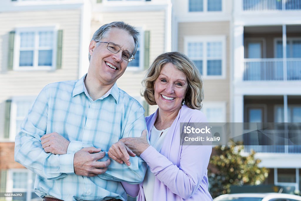 Mature couple in front of apartment building Portrait of a mature couple arm in arm, standing in front of an apartment building, smiling at the camera. 2015 Stock Photo