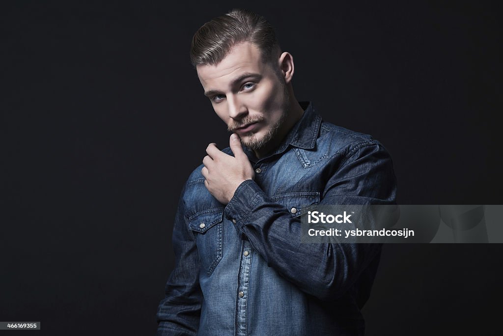 Casual winter fashion man. Wearing blue jeans shirt. Blonde hair. Casual winter fashion man. Wearing blue jeans shirt. Blonde hair and beard. Studio shot against dark wall. Adult Stock Photo