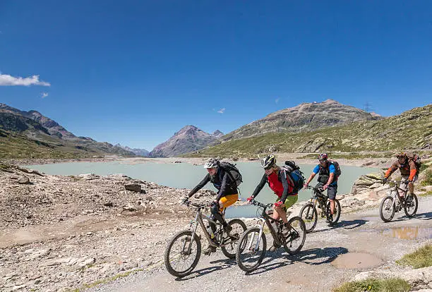 A Group of 4 mountainbikers is riding along the Lago Bianco ("White Lake") at an altitude of 2234 mt. (3730 ft.) on the Bernina Pass, Switzerland. It is a high mountain pass in the Bernina Range of the Alps in eastern Switzerland. It connects the famous resort town of St. Moritz in the Engadin valley with the Italian-speaking Val Poschiavo, which ends in the Italian town of Tirano in Valtellina.