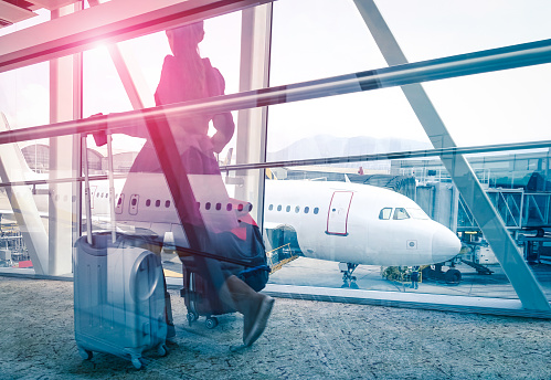 Travel concept with woman and suitcase moving fast to airport terminal gate - Double exposure look with focus on the aircraft in the background - Violet marsala sun flare with vintage filtered editing