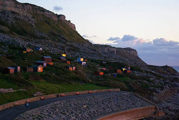 Beach-huts situated below the cliffs, above the promenade looking out to sea. The sun is going down causing a glow of orange across the huts highlighting their windows and benches on the prom below the huts. 