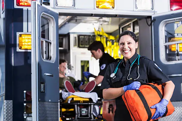 Hispanic female paramedic (30s).  Male paramedic helping boy inside ambulance behind her.