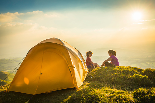 Two girls playing cards beside a bright yellow dome tent camped in an idyllic mountain top meadow illuminated by the warm light of a summer sunset. ProPhoto RGB profile for maximum color fidelity and gamut.