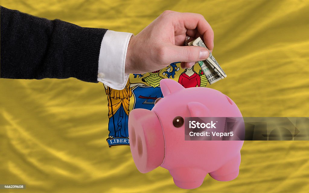 dollar into piggy coin bank and flag of new jersey Man putting dollar into piggy rich bank flag of us state of new jersey in foreign currency because of inflation 2015 Stock Photo