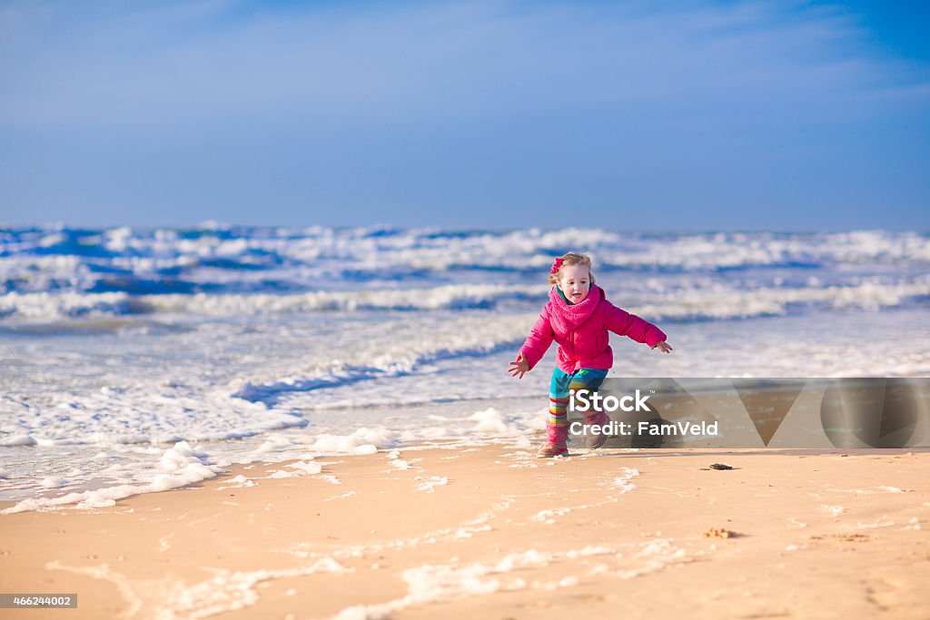 Little girl at a beach in winter Funny little girl, cute happy toddler in a colorful jacket, running and jumping on the beach at the North Sea in Holland, Netherlands, having fun on a sunny winter day 2015 Stock Photo