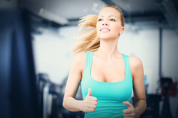 Attractive early 30's blond woman running on treadmill in gym. Wearing green tank top. Her hair is pulled back with a clip while her pony tail is in motion.