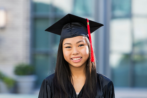 Portrait of an Asian teenage girl at her high school graduation, wearing a black cap and gown.