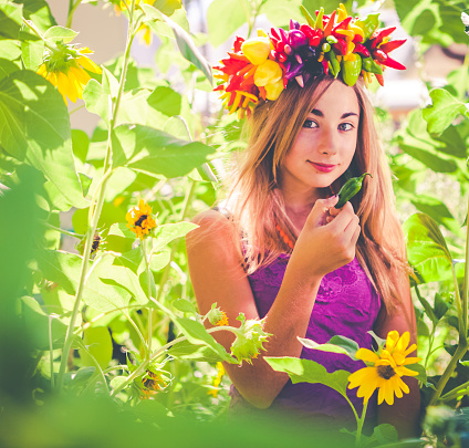 Long haired, blonde teen girl wearing a wreath of fresh peppers on her head, while in the garden. She's holding a green jalepeno pepper in her hand and smiling at the camera. It's a sunny summer day. Square composition.