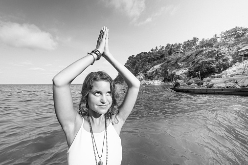 A radiant, young American woman in her 20s stands in a beach in Thailand. Her arms are raised overhead palms together in a yoga mudra. Surrounded by water she enjoys the present moment. Photographed with a Nikon D800.