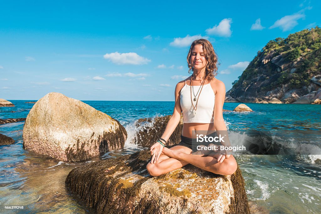 Meditating Woman Sits in Yoga Lotus Pose on Ocean Rock A radiant, young American woman in her 20s sits in Yoga Lotus Position on top of a rock in a Koh Phangan beach in Thailand. Surrounded by water she enjoys the present moment smiling while looking up. Her arms are stretched overhead. Photographed with a Nikon D800. 20-29 Years Stock Photo