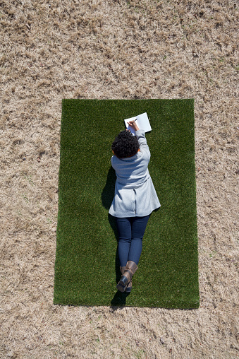 Aerial shot of a young woman laying on a small patch of green grass surrounded by a field of dead brown grass.  She is writing or drawing in a notebook.