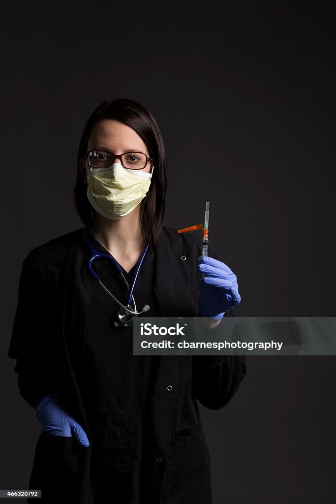 surgical mask wearing medical nurse with healthcare medicine vaccine syringe Upper body studio portrait of a caucasian medical nurse looking at camera with a stethoscope around her neck and holding a surgical syringe while wearing wearing scrubs, blue surgical gloves and yellow surgical mask. Canon EOS 5D Mark III. EF24-70mm f/2.8L USM. ISO 400, f/8.0 at 1/125th sec. 20-29 Years Stock Photo
