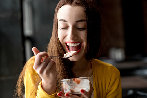 Young smiling woman dressed in yellow sweater eating ice cream in the dark cafe interior