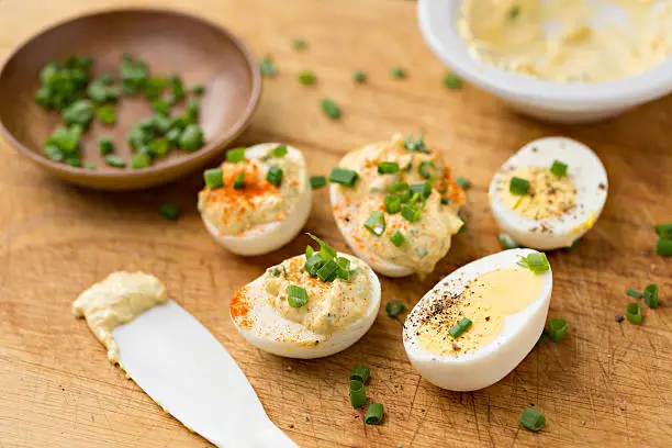 A high angle extreme close up horizontal photograph of a wooden cutting board with several assembled deviled eggs, a small plastic spatula, bowl of yolk mixture and a bowl with chopped scallions.