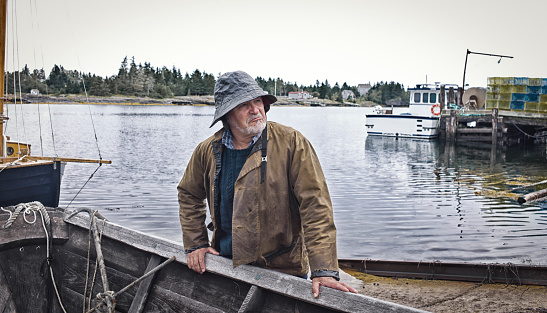 Fisherman pulling up a Dory on a slipway/wharf, Mahone Bay, Nova Scotia.  All gear and clothing is authentic to the era.  He has a southwester hat on and oilskin rain gear.  He is old with a white/grey beard.  Sea and sailboat make the background.  Leica camera photograph.
