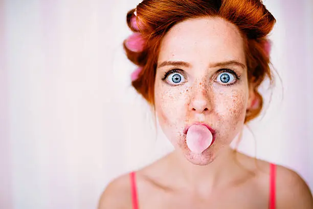 Photo of Young Redhead Woman With Curlers In Hair Blows Bubble