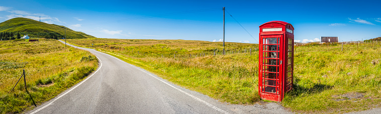 Iconic British red phone box deep in the picturesque rural landscape of the Scottish Highlands beside country lane and green hills under clear blue panoramic summer skies. ProPhoto RGB color profile for maximum color gamut.