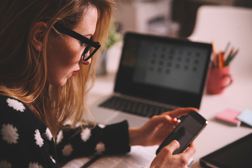 late night work in the office. portrait of a young blonde woman texting, using a smartphone in her creative office space.