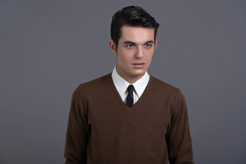 Retro 1950s fashion man with dark grease hair. Wearing brown sweater with black tie. Studio shot against grey.