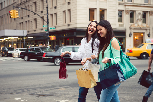 Two female friends walking in the street of New York City with shopping bags and smiling. 