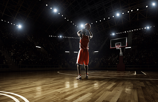 Basketball player in action on an indoor floodlit basketball court full of spectators.