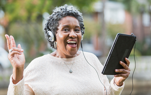 An excited senior, African American woman wearing headphones and using a digital tablet.  She could be listening to music, singing along.