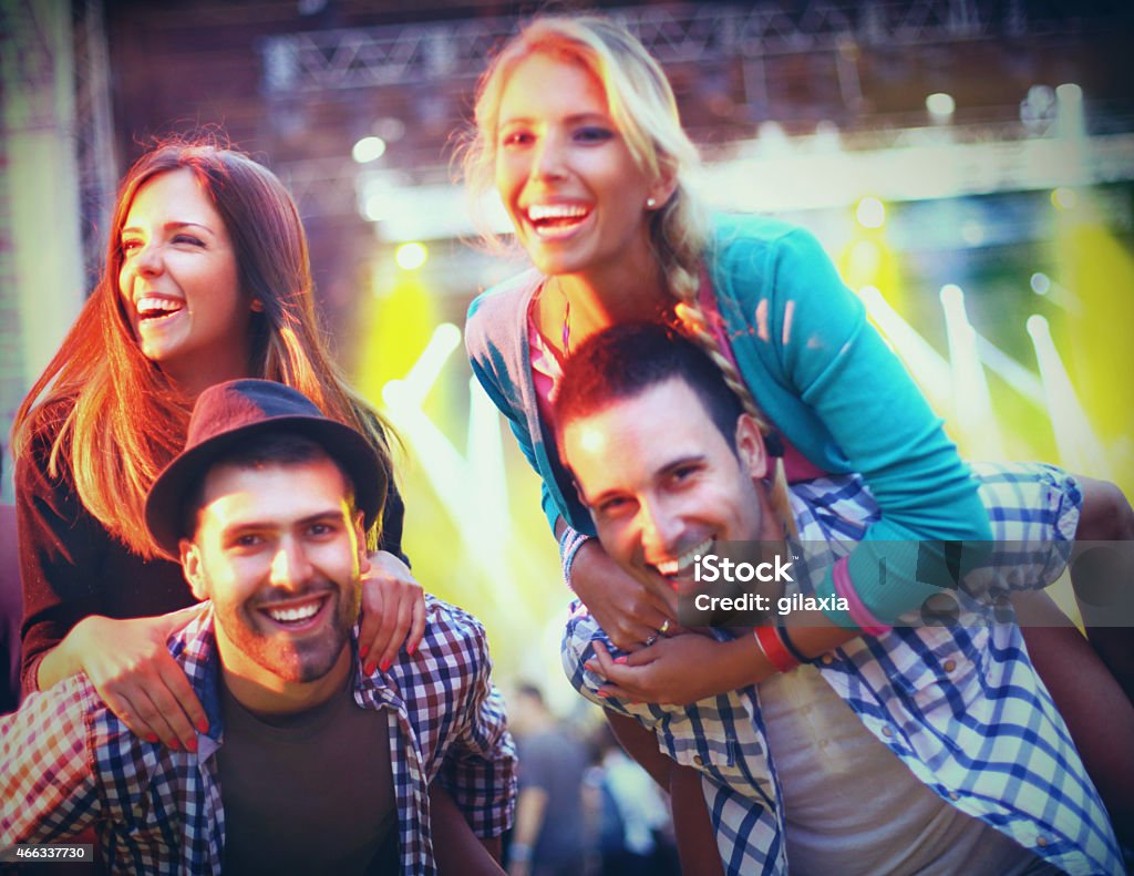 People enjoying a concert. Mid 20's people enjoying a concert.There two girls and two guys.Guys are piggybacking their girls. One girl is looking sideways.All looking at camera and laughing..Blurry stage in background toned purple green. 20-29 Years Stock Photo