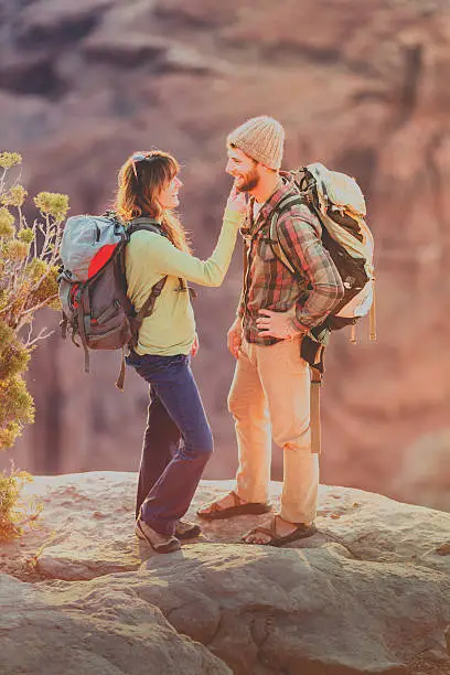 Cute couple interacting romantically with one another on a desert mountain hike near Moab, UT.  They are wearing trendy outdoor clothing and back packs she is flirtingly touching his face as they look at each other.  There is a sheer cliff next to them