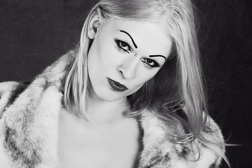 Horizontal B&W  studio shot on gray of young male-to-female transgender woman in fur coat.
