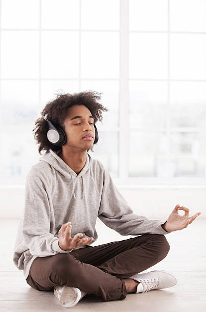 Chill time. Teenage African boy in headphones listening to the music and keeping eyes closed while sitting in lotus position on the floor teenage yoga stock pictures, royalty-free photos & images