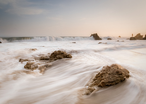 water rushes the shore at el matador beach in malibu california, creating imagery of classic california.  a man stands and watches the ocean far away on a pinnacle.