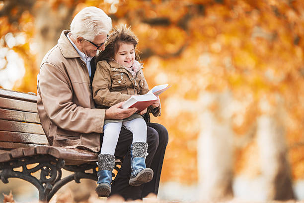 дедушка, читая книгу для детей девушка в парке. - grandparent grandfather granddaughter little girls стоковые фото и изображения