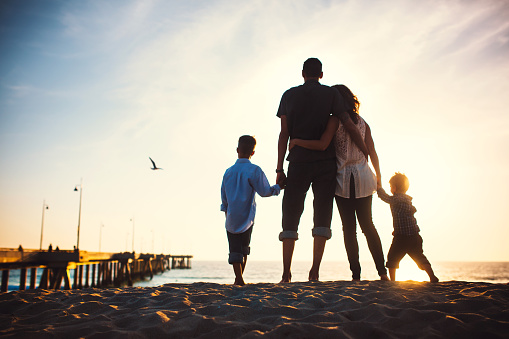 A silhouette of a beautiful young family at Venice beach, California watching the sun set over the ocean, the Venice Fishing Pier visible behind them.  The parents hug, holding the hands of their children.  Horizontal with copy space.