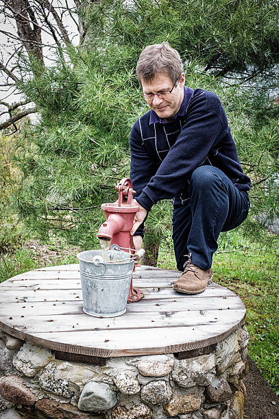 Mature Man Hand Pumping Water From a Well.  Hand pump is antique and red coloured.  Green trees and galvanized bucket.  Man is slim and wearing jeans and a sweater, both blue.  Rural Nova Scotia, Canada.   Leica Camera Photograph.