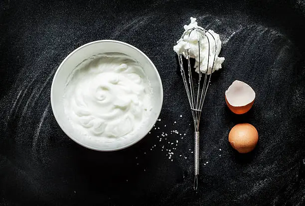 Whipped egg whites into a foam recipe - composition on black chalkboard from above. White bowl, eggbeater and eggshells.