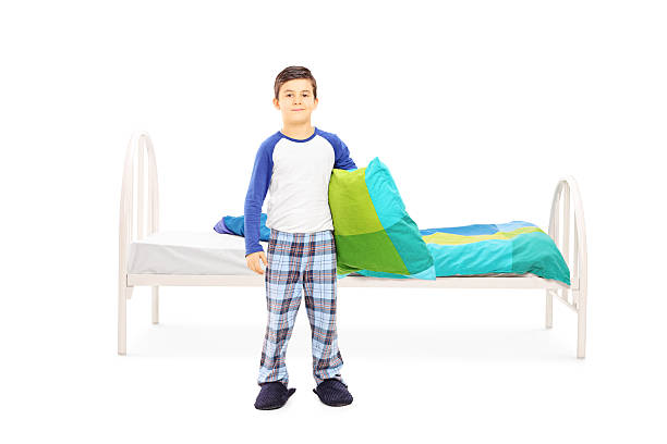 Full length portrait of a little boy with pillow standing in front of a bed isolated on white background
