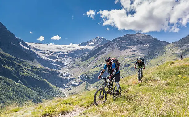 A couple of experienced mountainbiker is riding downhill nearby Alp Grum in front of the magnificent Palü Glacier situated in the Bernina Range in the canton of Graubünden in Switzerland.