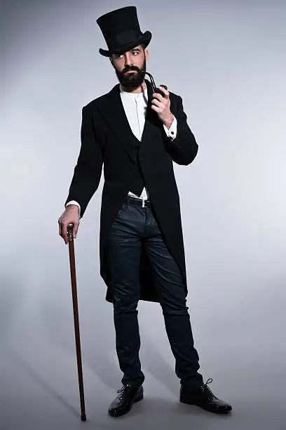 Retro hipster 1900 fashion man with black hair and beard. Wearing black hat. Standing with cane. Smoking pipe. Studio shot against grey.