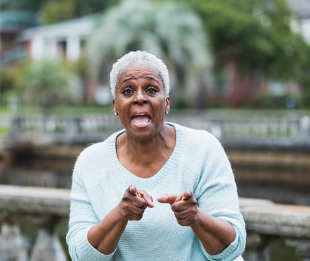 A senior, African American woman shouting at the camera with her mouth wide open, pointing with both fingers.