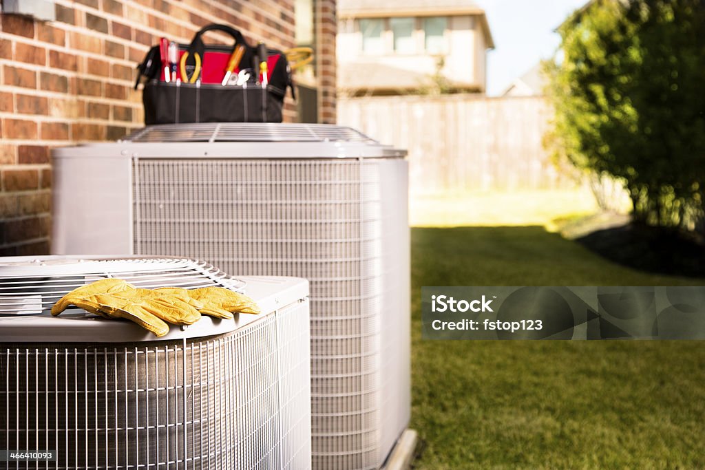 Service Industry:  Work tools on air conditioners. Outside residential home. Bag of repairman's work tools, gloves on top of air conditioner units outside a brick home.  Service industry, working class. Air Conditioner Stock Photo