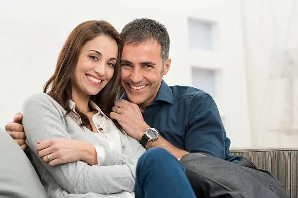 Happy Couple Embracing Sitting On Couch Looking At Camera