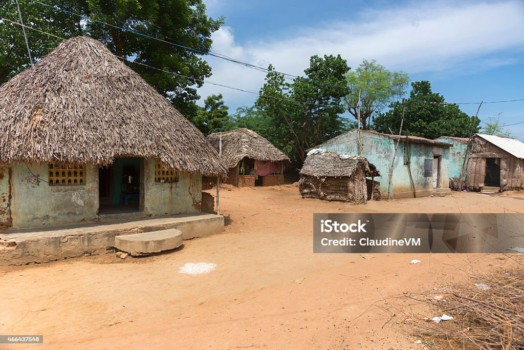 Group of humble dwellings in village. Kumbakonam, India - October 11, 2013: A group of humble houses, some with straw roofs stand at a dusty, sandy street against the background of green trees and a blue sky. Village Stock Photo