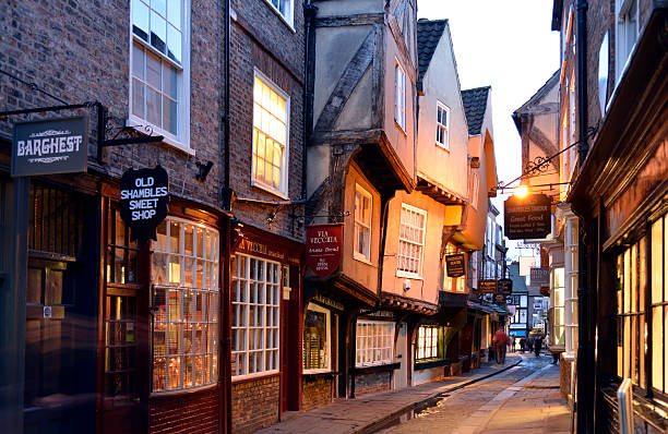 Shambles is probably the most famous street in York. Shambles has been a Shopping street since Tudor times and remains almost unchanged to this day and attracts tourists in the thousands.There are  people in the scene but none of them are recognisable due to a slow shutter speed. Although the sky is white it was almost the last light of the day. The street was very dark and the lights were coming on.