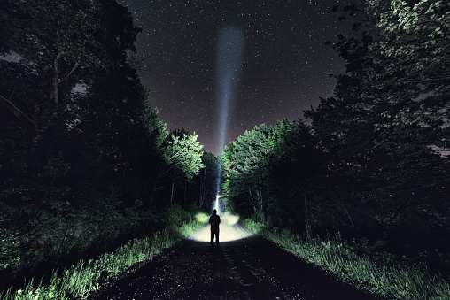 A man walks a narrow dirt road searching the skies with a flashlight.  Long exposure.