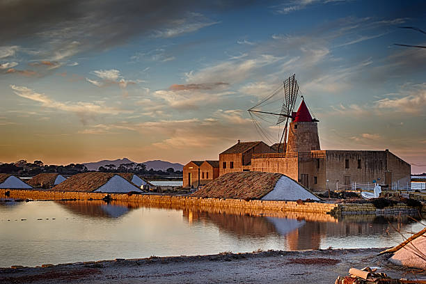 Salt flats mill A mill in "Stagnone" salt flats, between many salt heaps, mirror wather and marmalade sky.  Marsala, Sicily, Italy. salt flat stock pictures, royalty-free photos & images