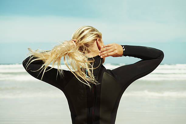 Time for a surf Rearview shot of a surfer girl tying her hair into a ponytail before hitting the water wetsuit stock pictures, royalty-free photos & images